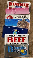 Stack of assorted novelty license plates including Ford Ronnie, breast cancer awareness, World's Greatest, cougar image, Florida Beef Council, Bonita Bay.