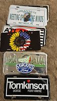 Stack of assorted license plates including Meeting of the Minds Key West, sunflower USA flag design, Sawgrass Ford, Tomkinson Dodge.
