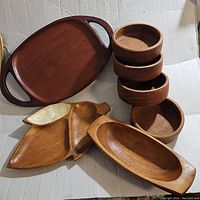 Photo showing a dark wooden serving tray with handles, stack of four round wooden bowls, a divided wooden serving tray with three compartments, and a narrow wooden serving bowl, all mid century style.