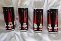 Four tall glasses displayed together, each with a black background panel and stylized red and white Santa Claus figures along with white star designs.