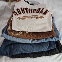 Photo of stacked clothing showing white Southpole T-shirt on top, brown patterned shirt, and multiple denim pants underneath