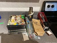 Overview of the entire lot on kitchen counter showing all items including the metal tin, martini shaker, bags of swizzle sticks, drip collar in box, and paper bag labeled 'Amigo Collection'.