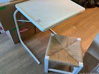 Photo showing white adjustable bed table and small rush seated stool on wood floor with boxes in background.