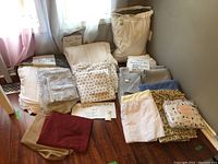 Photo showing multiple bedding pieces including sheets, pillowcases, blankets, and comforters stacked on the floor labeled with notes for twin and queen bedding.