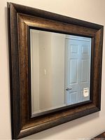 Framed beveled edge square mirror hanging on a wall, showing the dark textured wooden frame and clear reflective glass with beveled edges.