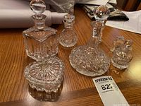 Five clear crystal items arranged on a wooden table including decanters, a heart-shaped ring dish, and a vinegarette.