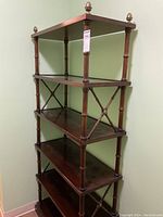 Tall dark wood shelving unit with six shelves and brass finials on top corners, positioned against a green wall.