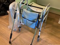 Photo showing aluminum folding walker with blue fabric pouch on wooden floor.