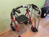 Red folding walker with padded seat, black handles with brakes, and four wheels beside a silver folding walker and silver folding cane all placed on wooden floor.