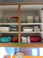 Overview of kitchen cabinet shelf showing assortment of glass casseroles, ceramic baking dishes, metal casserole pots in turquoise and red, and three small round patterned storage containers.