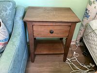 Full view of the wooden night stand showing drawer and lower shelf, positioned next to a light blue upholstered chair.