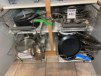 Photo shows kitchen cabinet pull-out shelves with various pots and pans mostly in stainless steel and black non-stick finishes. Several have glass lids and metal handles. Includes a small saucepan and frying pans with blue and metal handles.