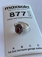 Silver ring with red oval stone set within a geometric design accented with diamond-like chips, photographed on auction note card.