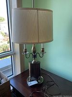Full view of table lamp showing brass base, two light sockets, beige fabric shade, and tall brass finial standing by window