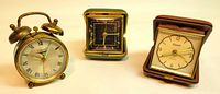 Lot of three vintage travel clocks shown side by side: Waltham twin bell brass alarm clock, Fisher folding travel clock with black face, Forestville folding clock with cream face