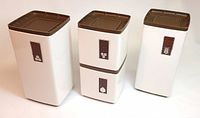 Four metal kitchen canisters grouped. White bodies with brown textured vertical tabs and silver icons. Brown plastic lids. All vintage with some wear.