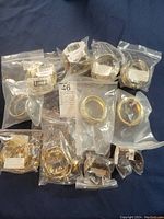 Overview photo showing 11 assorted gold-tone bracelets individually sealed in clear plastic resealable bags with labels and barcodes, arranged on dark blue fabric.