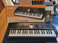 Overview of Casio SA-76 electronic keyboard next to original orange box showing the product image and model number