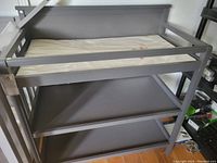 Front angled view showing gray wooden changing table with two lower shelves and padded changing mat on top.
