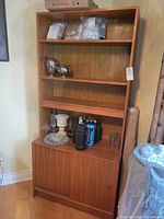 Full view of teak cabinet showing three open shelves with various items placed on them and sliding door compartment at the bottom.