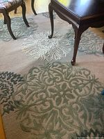 Close-up side view of the area rug under a wood coffee table and a chair with floral upholstery, showing the beige base color and raised floral patterns in green and cream tones.