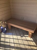 Wooden slatted bench and blue glass vase with flowers placed on a wooden deck next to wooden siding wall, captured from the front and side angles.