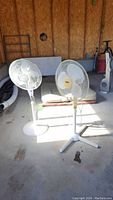 Two white pedestal fans standing on a concrete floor with other items in the background, untested condition.