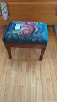 Wooden footrest with floral needlepoint upholstered cushioned top shown from a front angle revealing overall condition and structure.