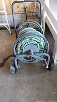 Photo shows heavy duty metal caddie with green 50 ft garden hose coiled around it, viewed from front. Metal frame and wheels visible.