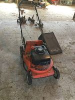 Frontal view of orange Ariens gas powered lawn mower with attached black fabric grass bagger inside an indoor workspace with concrete floor.