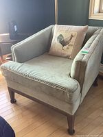 Front and side view of upholstered mid century chair with light green decorative patterned fabric and wood trim. Includes a pillow with a rooster print.