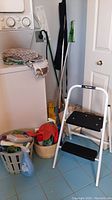 View of laundry area showing Swiffer mop, step ladder, bucket of cleaning sprays and detergents on blue tiled floor, mop and folded linen on washing machine