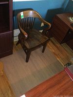 Side angle of wooden office chair on clear plastic mat showing seat, armrest details, and floor protection mat.