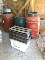 Lot overview showing two red rain barrels, one green barrel, vintage wooden crate on top of white styrofoam cooler.
