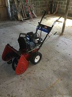 Three-quarter front view showing red two-stage auger housing, black plastic chute and handle assembly