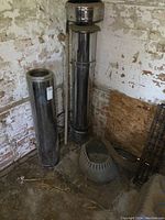 Full view of both stovepipe sections and roof flashing placed on dirt floor in a corner with old white painted brick walls.