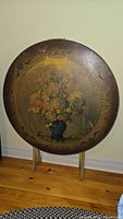 Full view of the vintage round tilt-top table standing upright showing floral painting on tabletop.