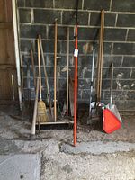 Photo showing pick axe, shovels, pole saw, rakes, broom, and red yield sign leaning against stone wall in barn or shed