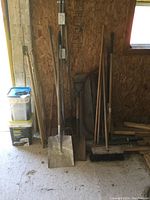 Photo showing assortment of wood handle yard and shop tools including shovels, pitchfork, broom, and buckets of ice melt.