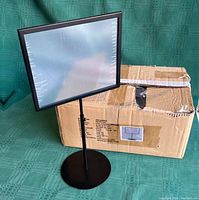 Six adjustable black sign holders shown, three assembled and three disassembled with components including square frames and round bases, displayed with original cardboard box.