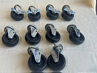 Ten black rubber caster wheels arranged on a light-colored fabric surface. Eight wheels have metal mounting plates, and two wheels have threaded stems for attachment. Wheels are 4 inches in diameter.