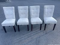 Front view of four beige upholstered dining chairs with button tufting, dark wooden legs, arranged in a row outdoors on an asphalt surface.