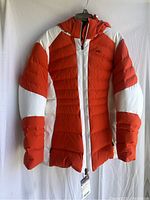Front view of the Kjus Duana orange and white two-tone down jacket hung on a hanger showing the full front and attached hood.