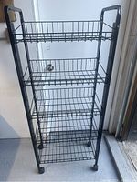 Front view of black metal shelving cart with 5 wire mesh shelves and caster wheels in lighted room.