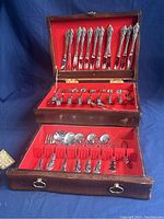 Open wooden chest with hinged lid and pull-out drawer, lined in red velvet, holding vintage Oneida flatware with decorative handles.