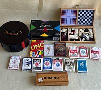 Photo showing all game and playing card items laid out including various decks of cards, Trivial Pursuit box, domino set, chess/checkers set and round black case.