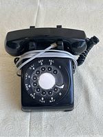 Top front view of the black rotary dial phone showing the handset, rotary dial, and cords.