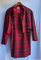 Two-piece vintage women's suit by Albert Nipon in red plaid wool fabric, including jacket and dress, hanging on wooden hangers against a white backdrop.