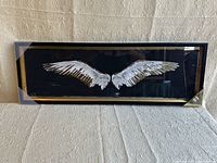 Wall art with eagle wings design in glass front wood frame against textured beige wall.