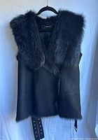 Front view of black lamb leather sleeveless vest with large dark fur collar and fur lining visible at edges, hanging on hanger against white background.